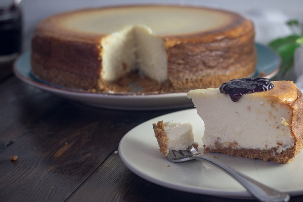 "Cream Cheese Brownies: A sliced creamy cheesecake on a plate with a whole cheesecake in the background, showcasing a rich, golden crust and topped with a dollop of dark berry compote.